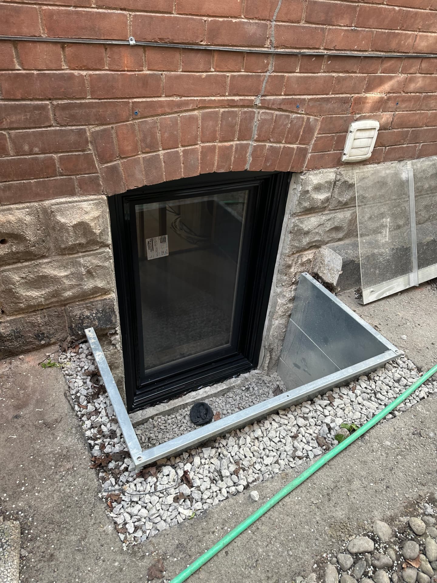 Black-framed inswing basement egress window set into a brick and stone foundation with a galvanized window well and gravel