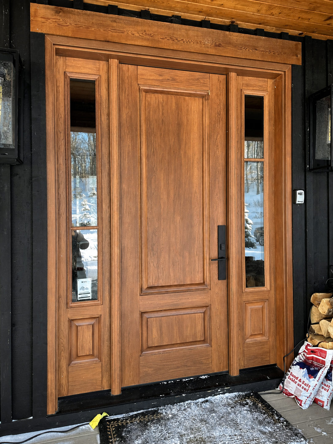 Wood-grain front entry door with a raised panel, flanked by two tall glazed sidelites, set in black vertical siding under a timber header