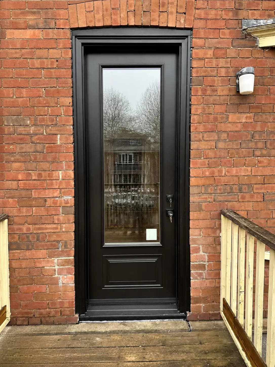 Black entry door with a tall clear glass lite over a raised bottom panel, set in a red brick wall above a wood deck