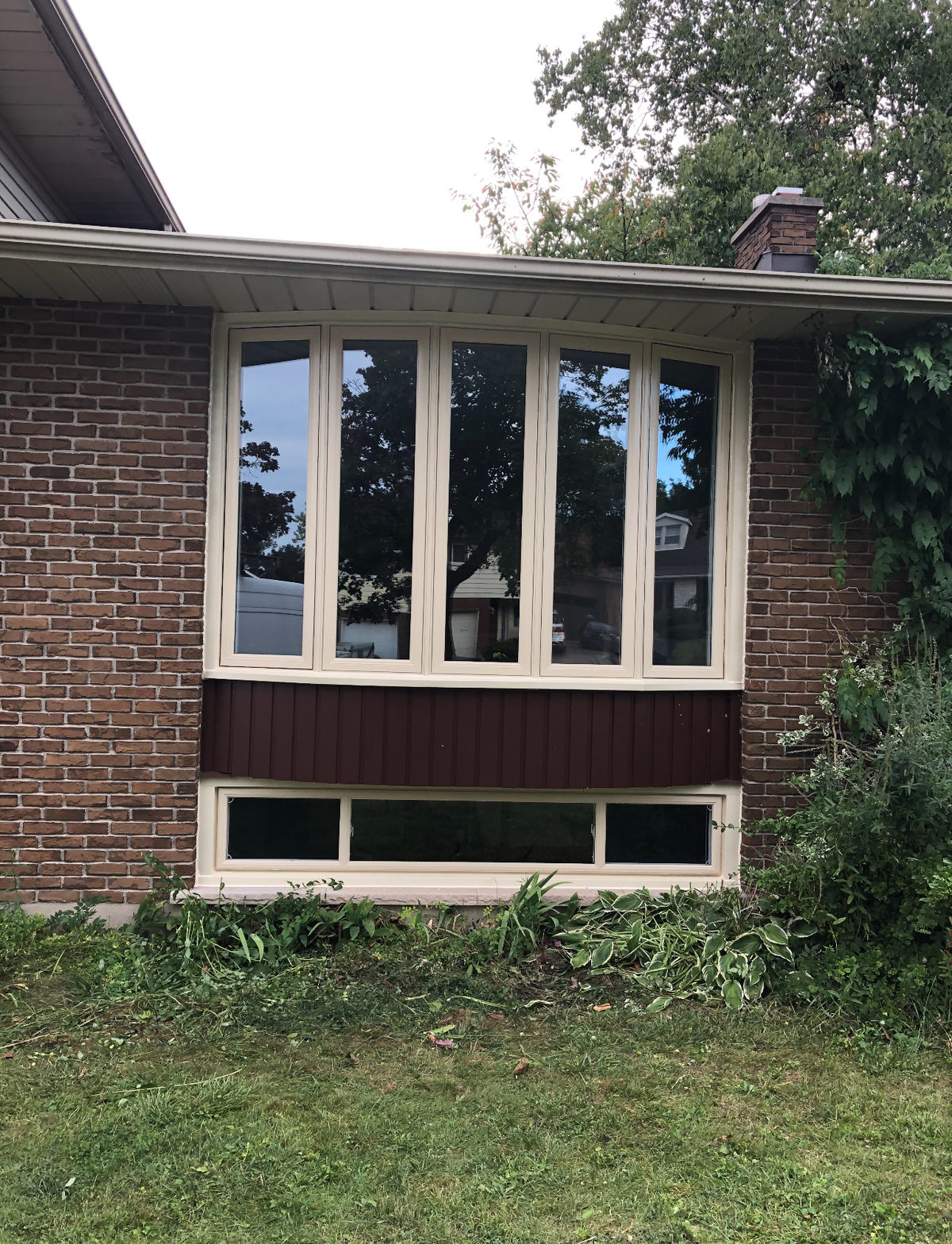 Five-panel cream-framed bow window in a brick wall above dark red cladding, with a long low basement window underneath