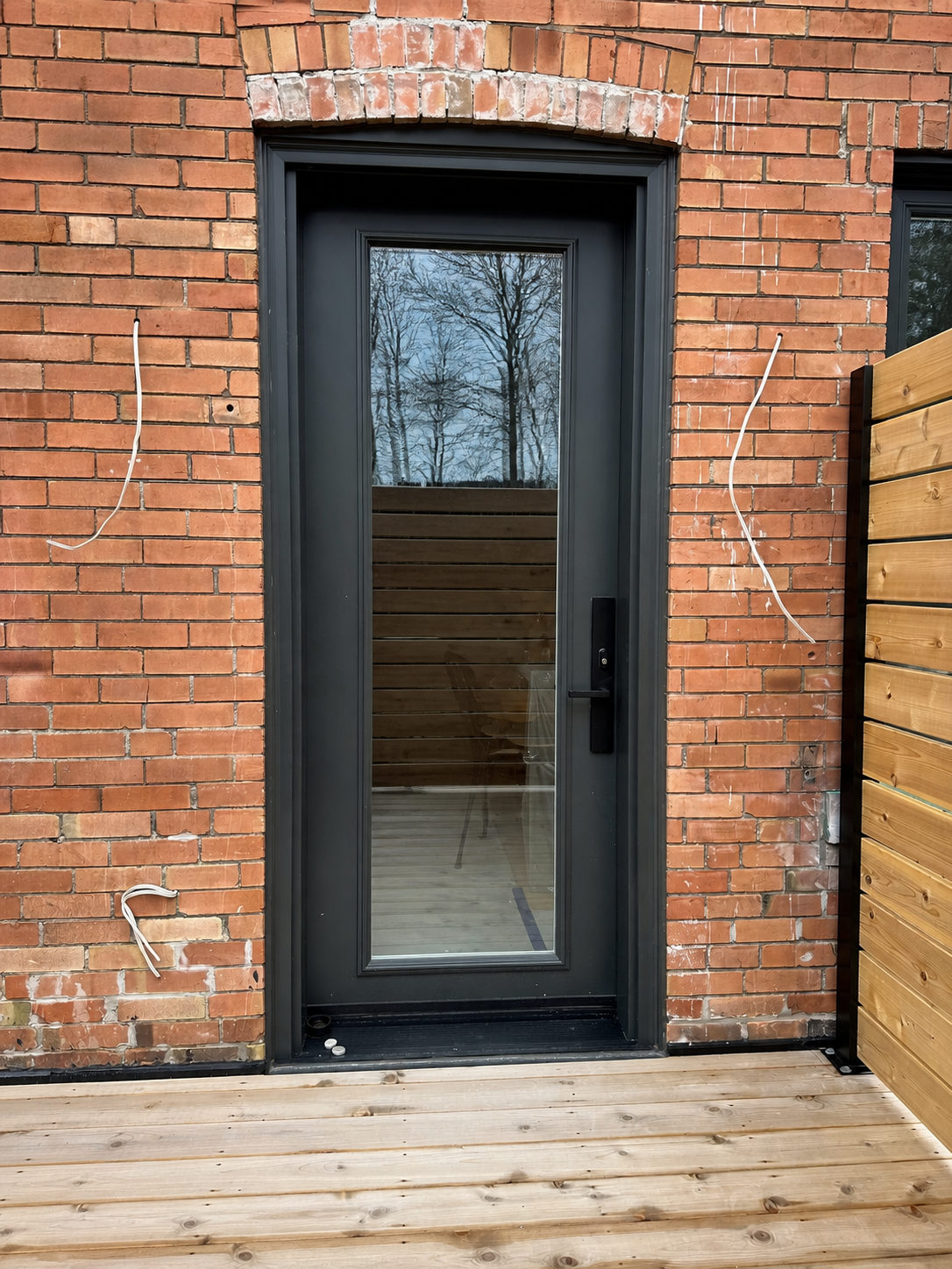 Black full-glass entry door with a black frame set into a red brick wall with an arched brick header, opening onto a cedar deck