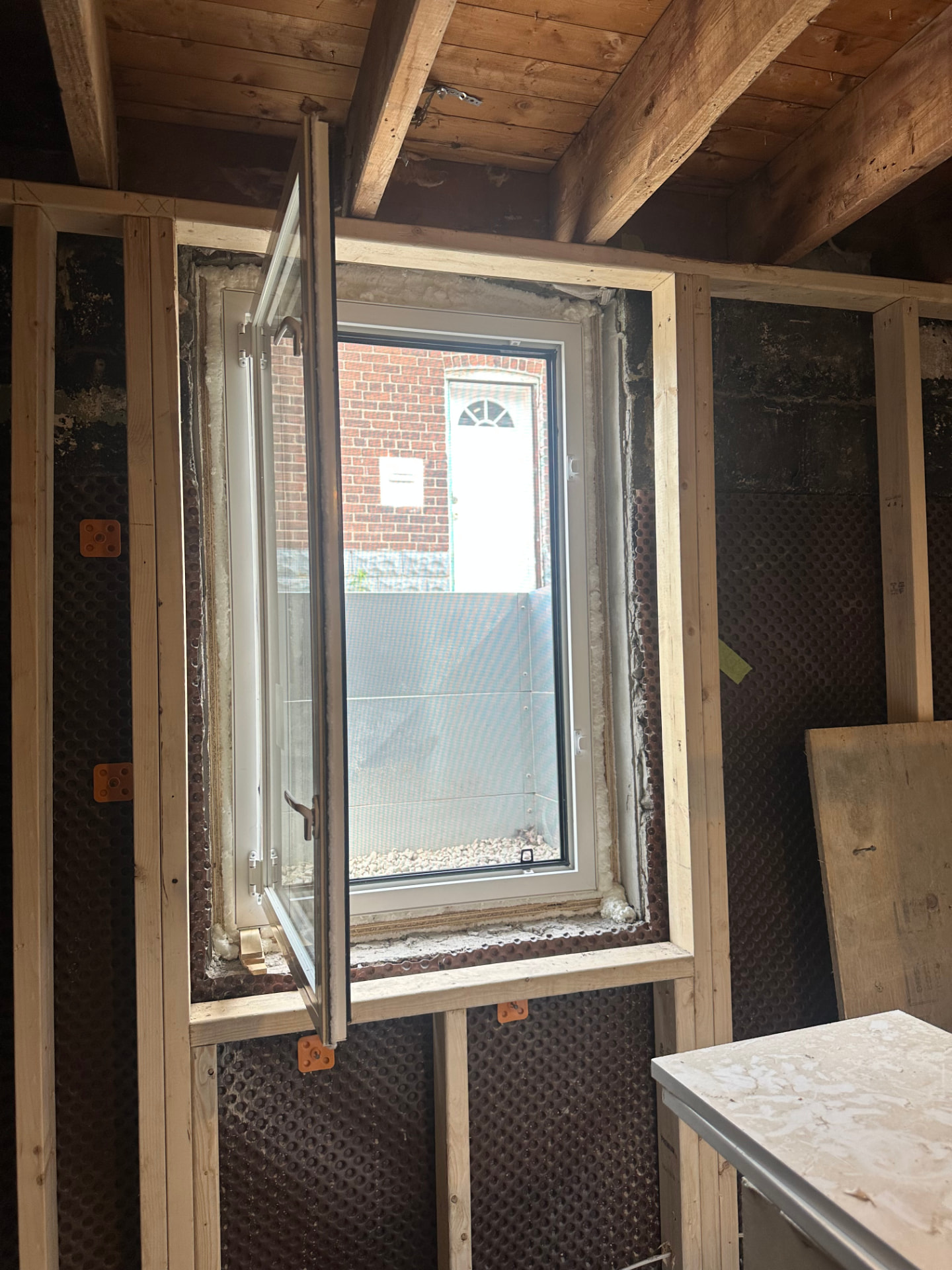 Interior basement view of the same egress window swung inward on its hinges, in a stud-framed opening with spray foam around the frame