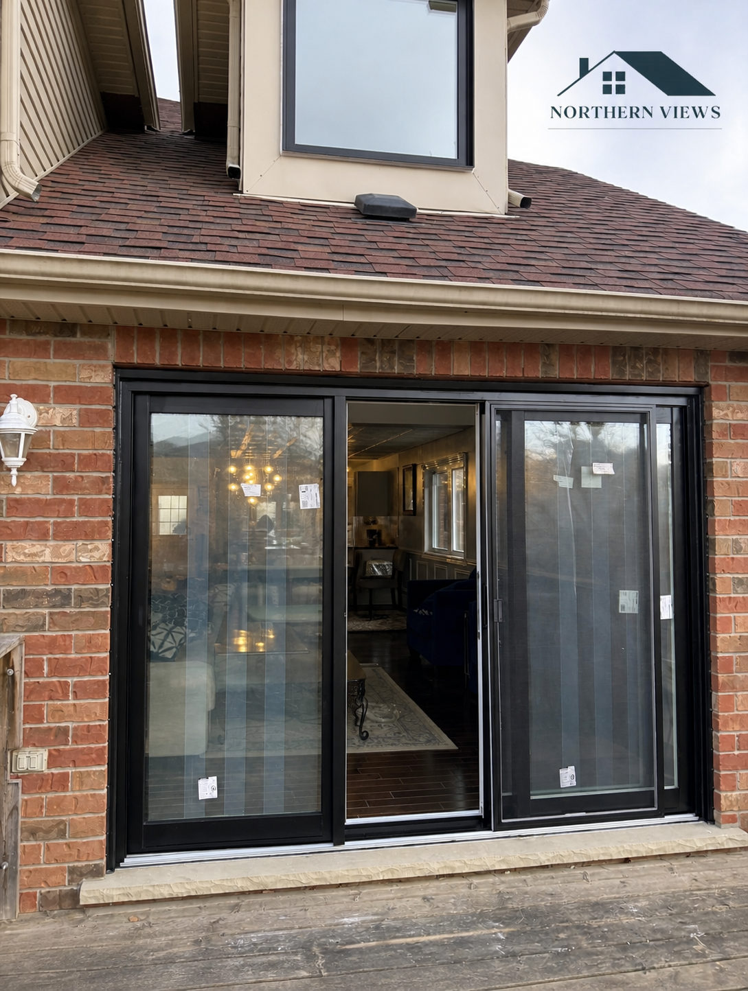 Three-panel black sliding patio door with a screen panel, installed in a red brick wall opening onto a wood deck