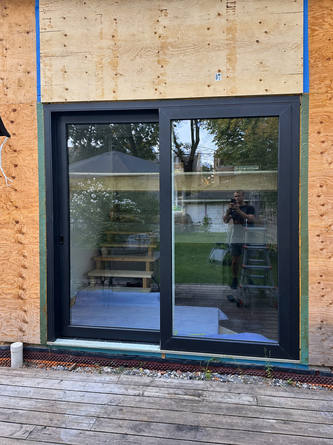 Black two-panel sliding patio door installed in a plywood-sheathed wall, with green flashing tape around the frame and a wood deck below
