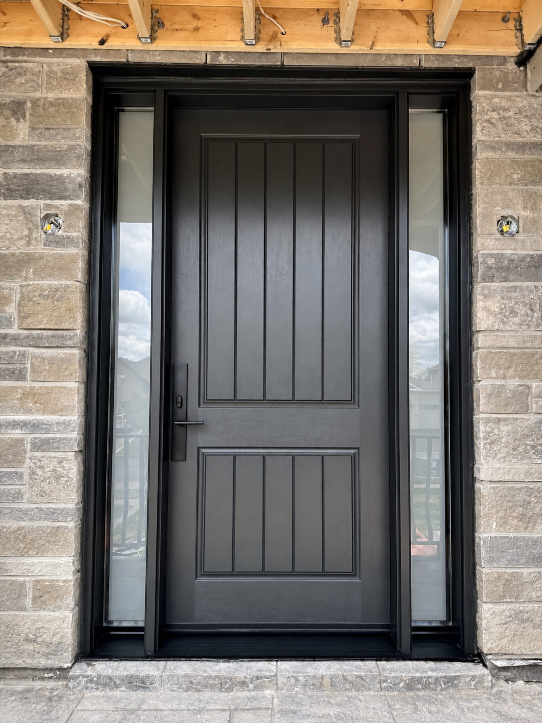 Black two-panel plank-style entry door with a black handleset, flanked by two tall clear glass sidelites in a light stone wall