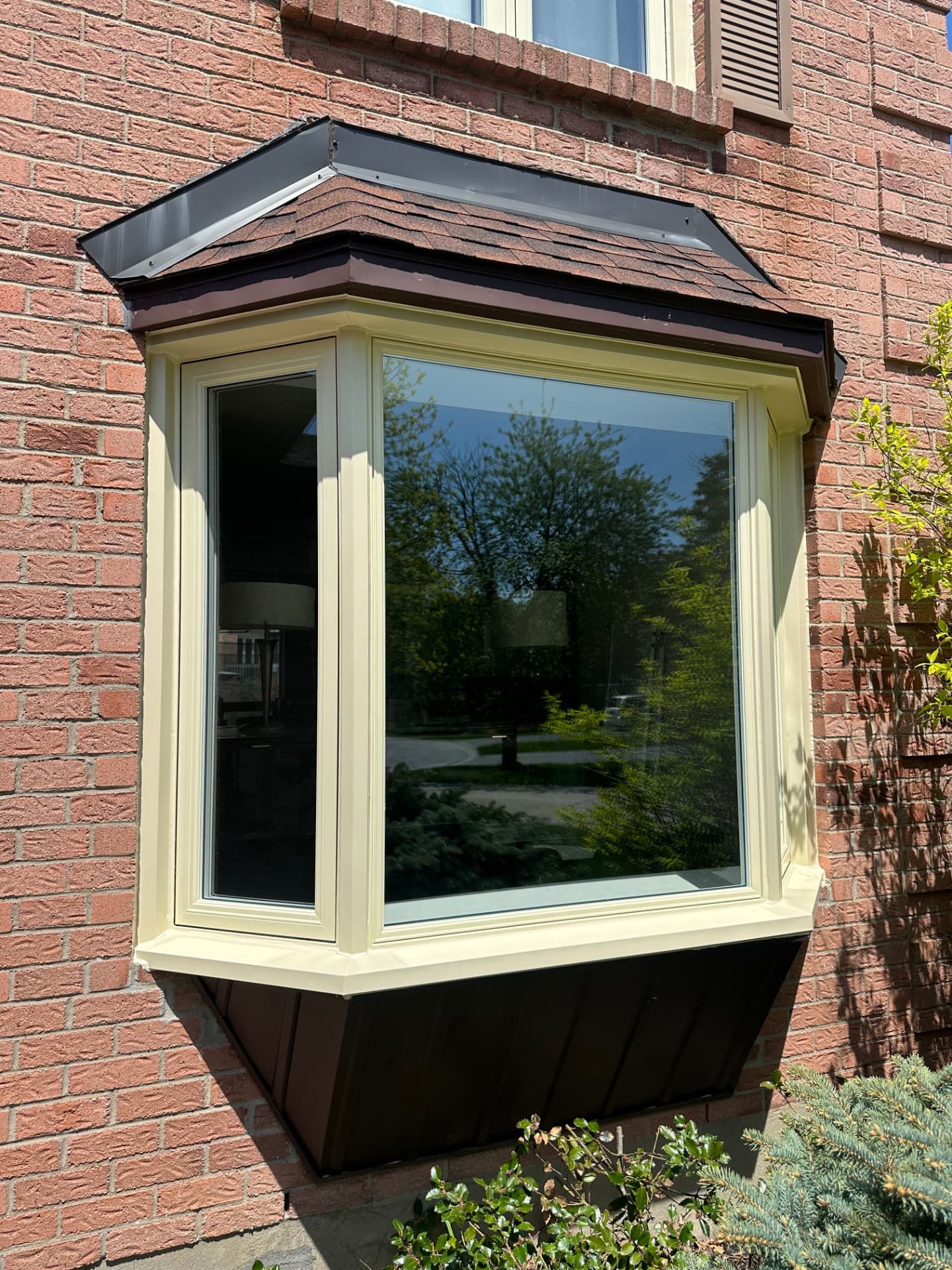 Cream-framed bay window projecting from a red brick wall with a shingled roof cap and dark brown cladding below