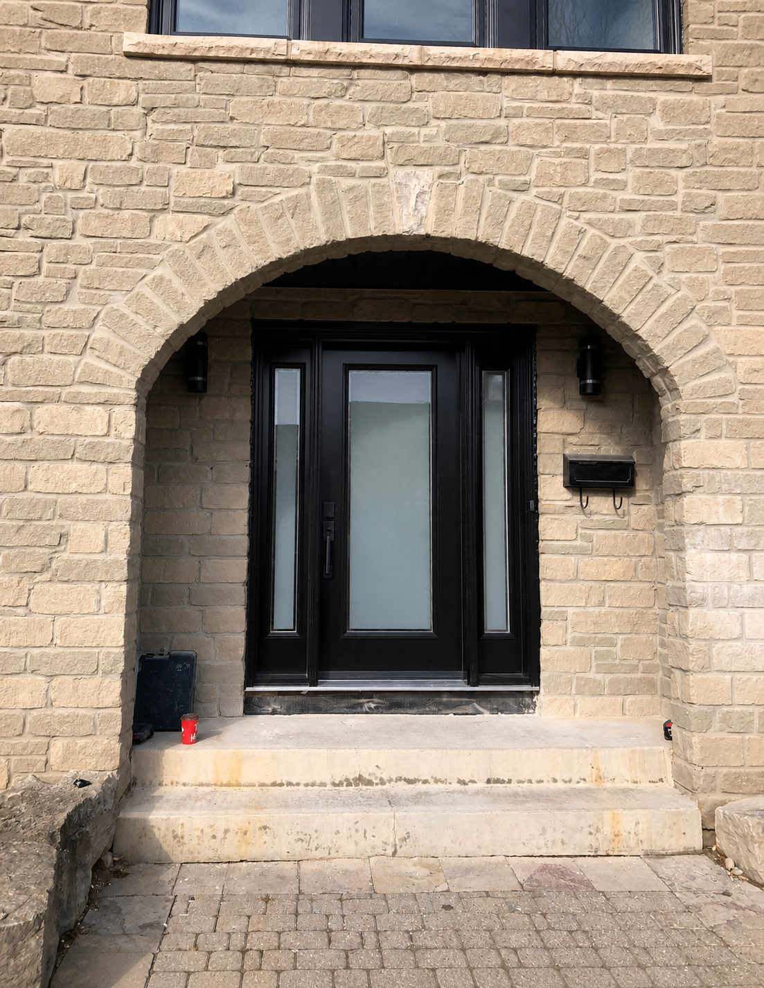 Black front entry door with a frosted glass panel and two frosted sidelites, set beneath a beige stone arch
